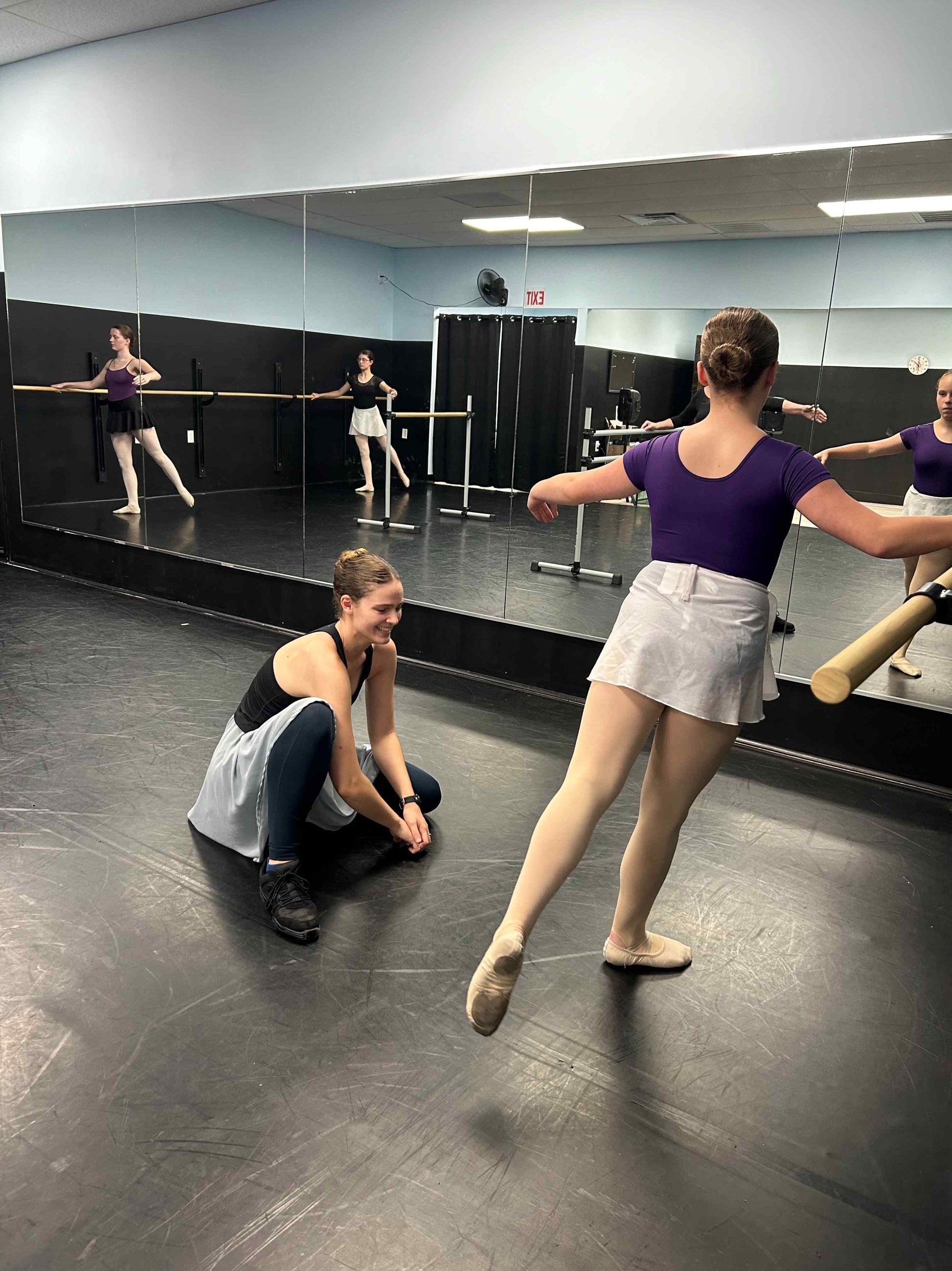 Ballet class with multiple dancers at the barre, teacher correcting a student on the floor