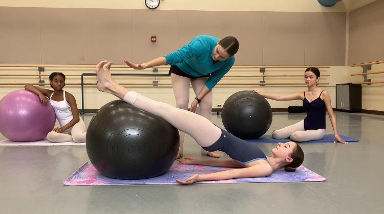Ballet student corrected personally on demi-pointe alignment while completing PBT exercise on yoga ball. Two other students observe.