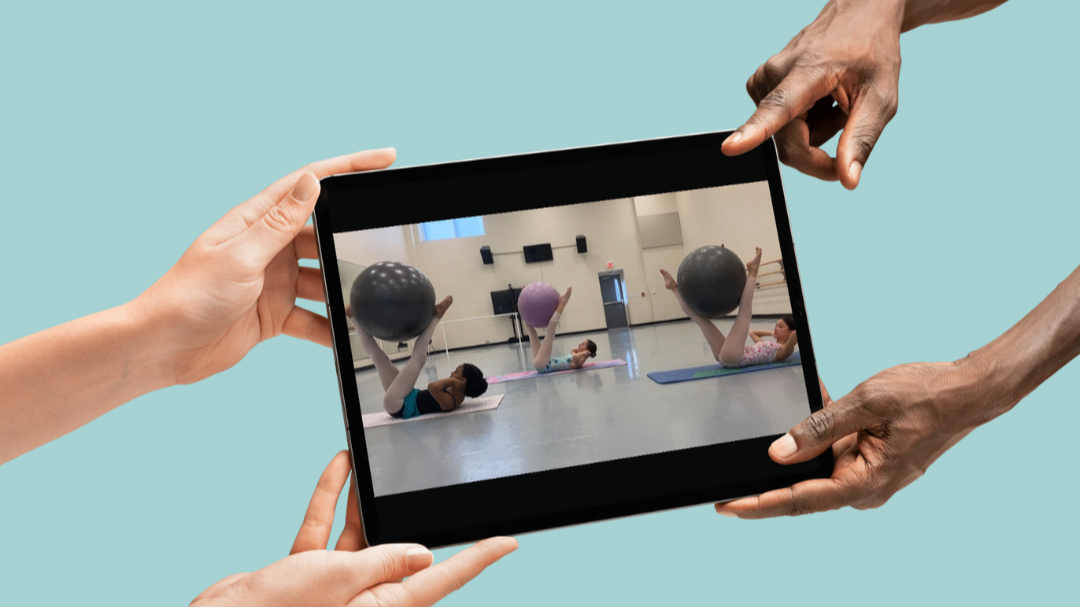 Group PBT class with three ballet students performing exercise with yoga ball. Image shown on tablet held by two sets of hands with light teal background.